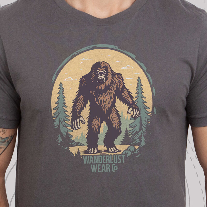 Model wearing Wilderness Watcher graphic tee featuring full-color Bigfoot illustration framed by pine trees and a vintage sunset backdrop